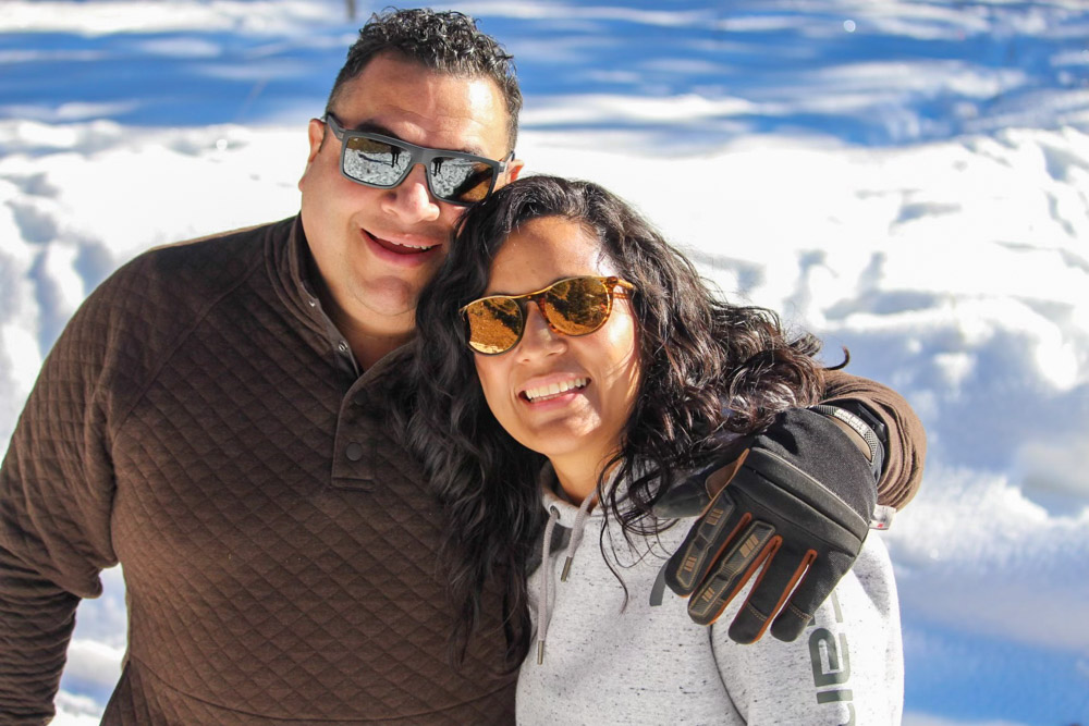A smiling couple posing together in a snowy landscape, both wearing sunglasses and winter attire. The man has his arm around the woman, and they are standing close, with snow-covered ground and bright blue sky in the background.