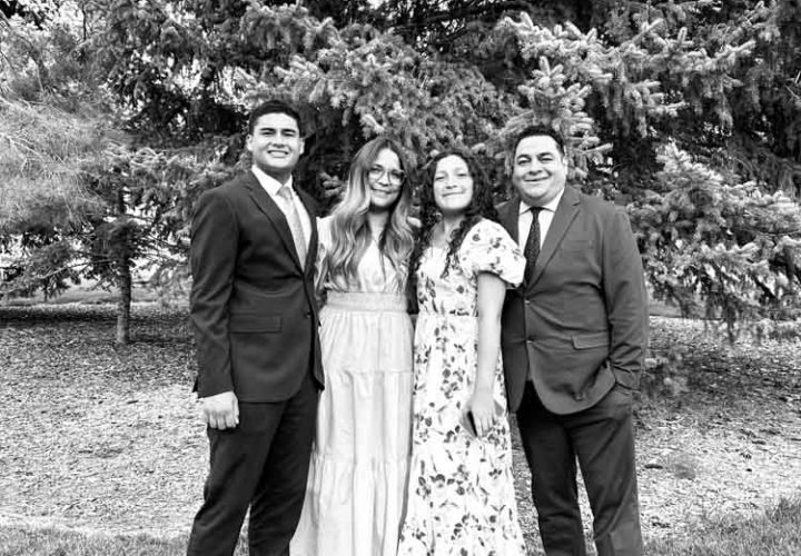 A black-and-white photograph of four adults standing together outdoors in front of evergreen trees. Two men are dressed in suits, and two women wear long dresses. They are smiling and appear to be posing for a formal family photo on grassy ground.