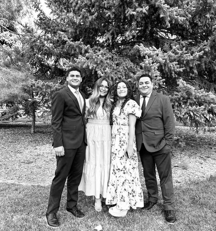A black-and-white photograph of four adults standing together outdoors in front of evergreen trees. Two men are dressed in suits, and two women wear long dresses. They are smiling and appear to be posing for a formal family photo on grassy ground.